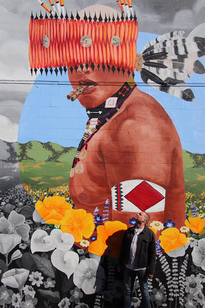 David Dowd stand at the foot of a street art mural depicting a Hopi native amidst California hills and poppies